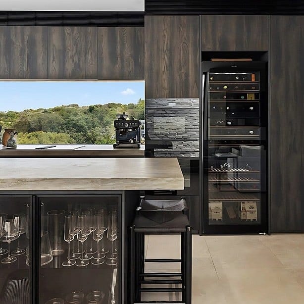 Dark timber kitchen with a stone-topped island, glass-fronted cabinets and a built-in wine fridge beside a window framing bushland