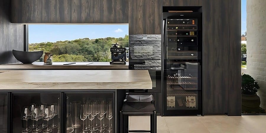 Dark timber kitchen with a stone-topped island, glass-fronted cabinets and a built-in wine fridge beside a window framing bushland