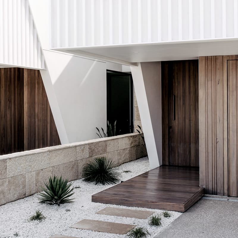 Timber pivot door and floating timber step at the Luke Parker duplex, beneath a white batten-screened upper level, with an angled white column and gravel garden