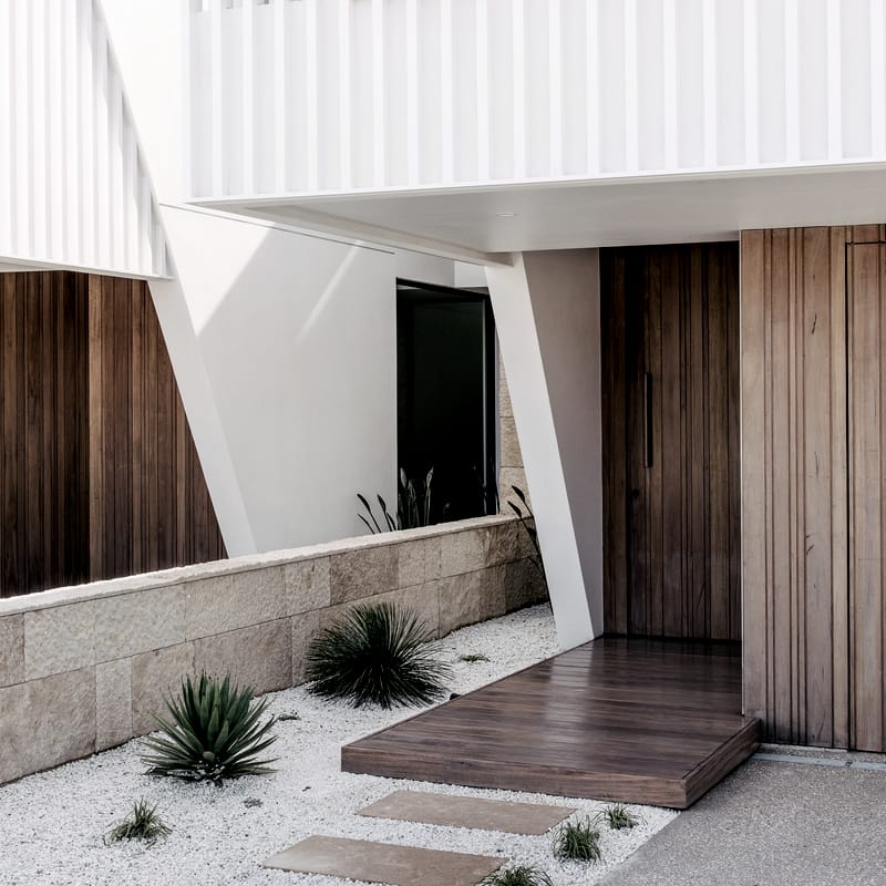 Timber pivot door and floating timber step at the Luke Parker duplex, beneath a white batten-screened upper level, with an angled white column and gravel garden