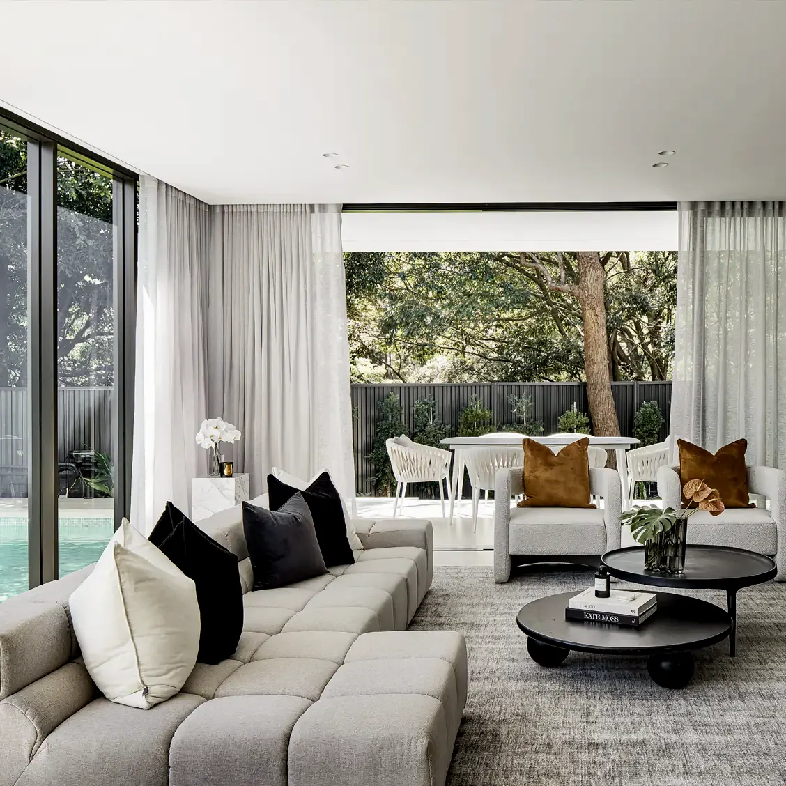 Living room with a modular sofa and round coffee tables, opening through glass to a garden and pool