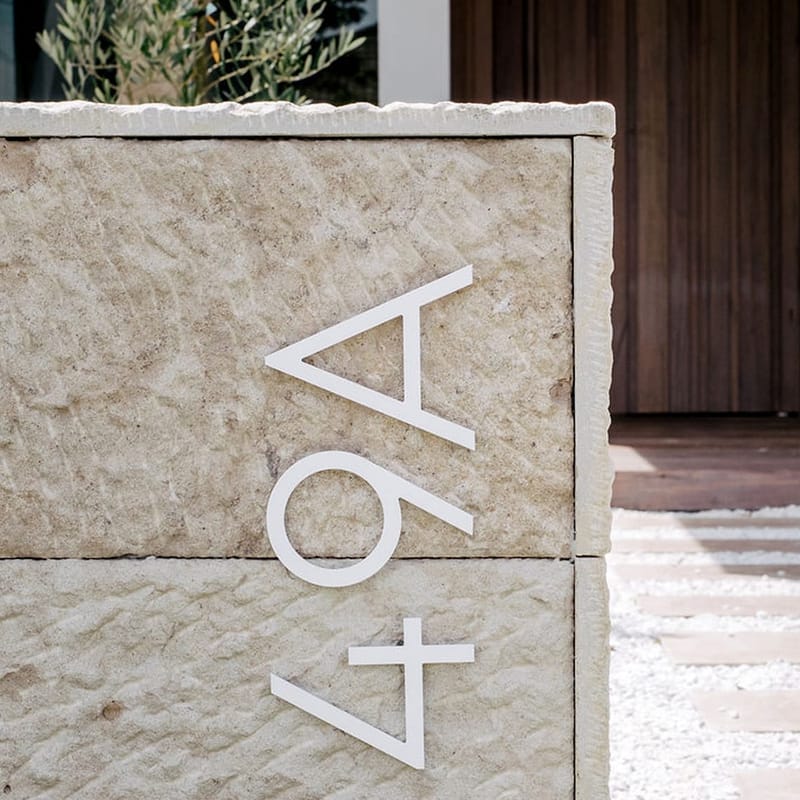 Close-up of a hand-chiselled sandstone wall with white metal house numbers, a white gravel path and a timber entry behind