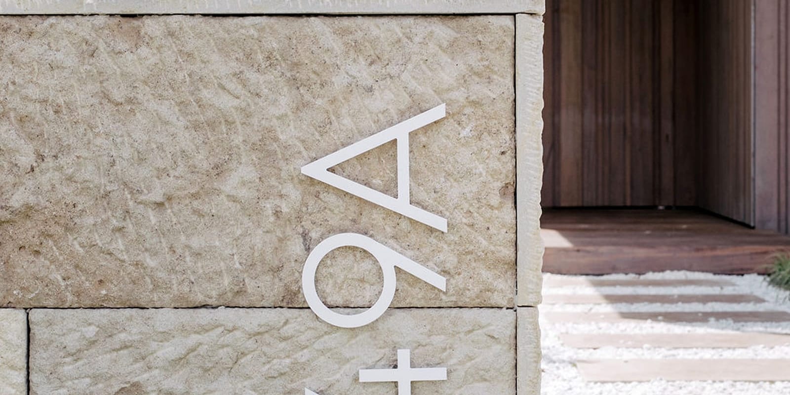 Close-up of a hand-chiselled sandstone wall with white metal house numbers, a white gravel path and a timber entry behind