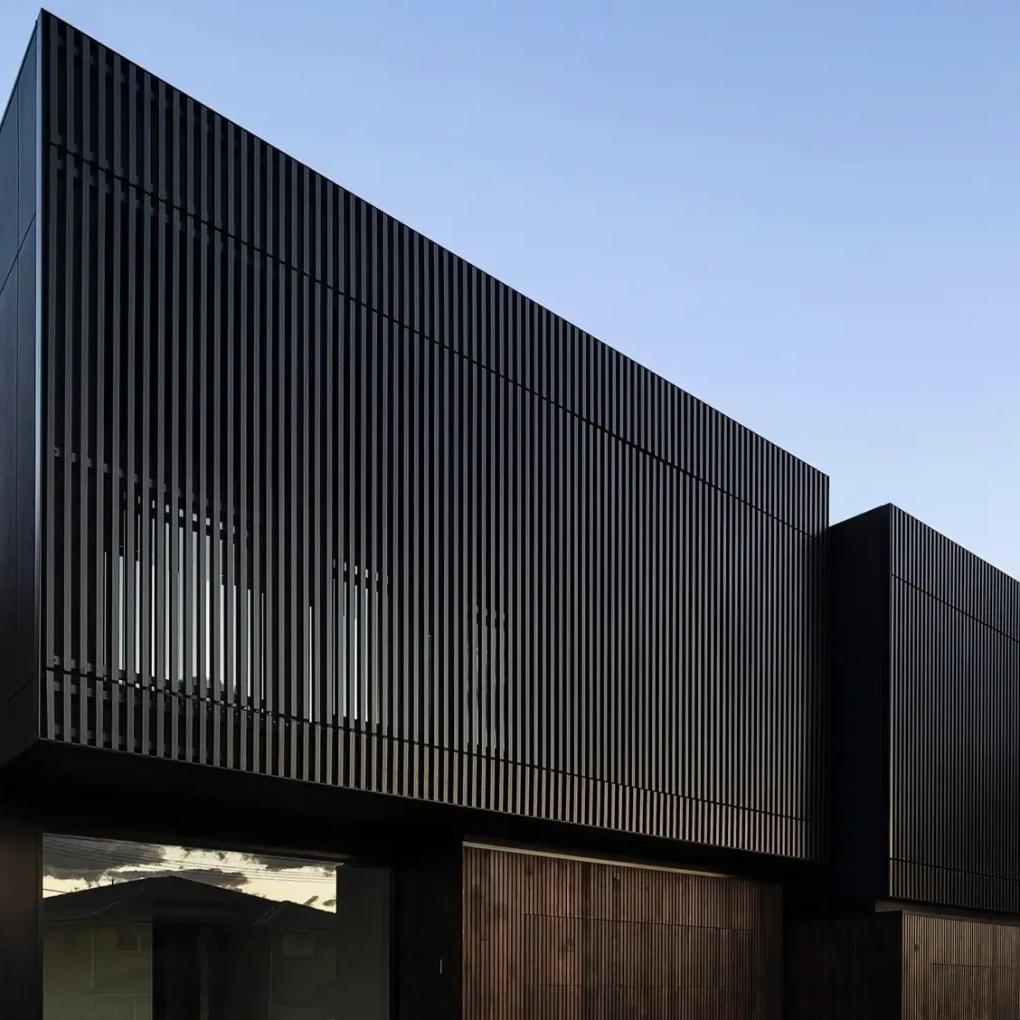 Duplex at dusk with an upper floor clad in dark vertical battens floating over timber-clad and glazed ground-floor rooms