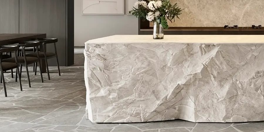 Kitchen with a rough-hewn stone island bench, crazy-paved stone floor, dark timber joinery and a dining table beyond