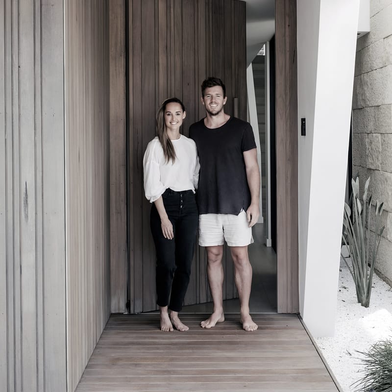 Luke Parker standing barefoot in a timber-lined entry beside a tall Duplex timber door, white render and a stone wall with coastal planting