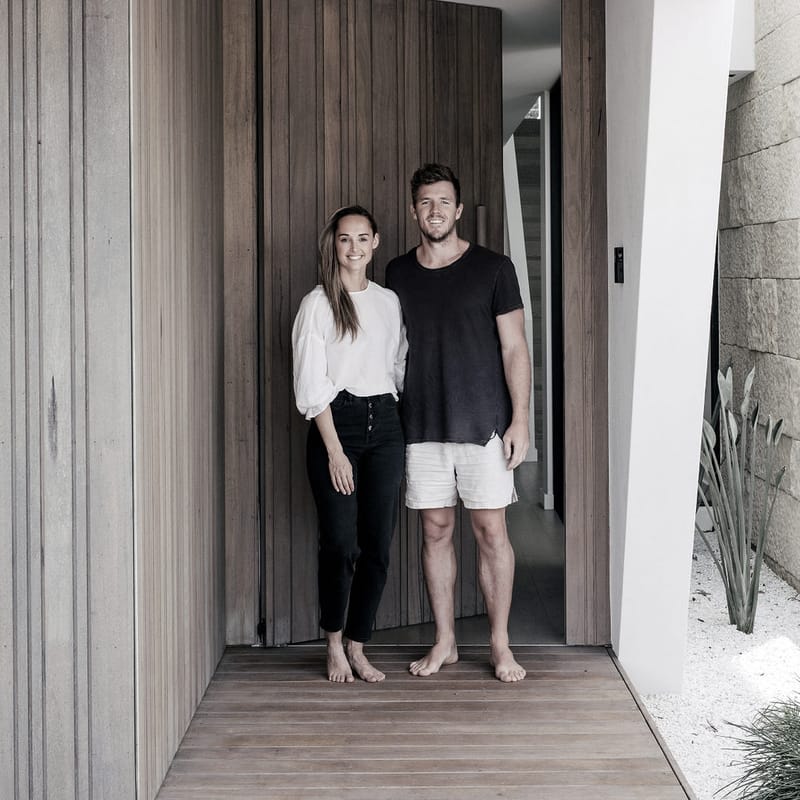 Luke Parker standing barefoot in a timber-lined entry beside a tall Duplex timber door, white render and a stone wall with coastal planting