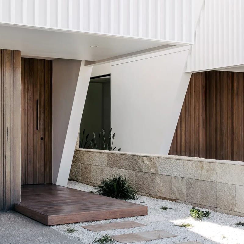 Recessed timber entry with a timber step and angled white column beside a sandstone garden wall, under a white batten-screened upper level