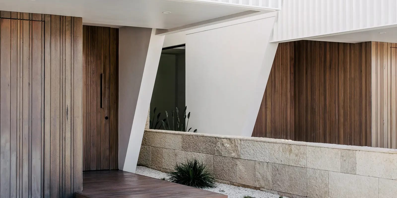 Recessed timber entry with a timber step and angled white column beside a sandstone garden wall, under a white batten-screened upper level