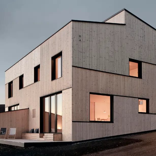 Two-storey house clad in pale vertical timber with deep-set black-framed windows glowing at dusk