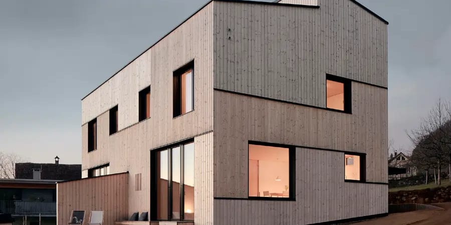 Two-storey house clad in pale vertical timber with deep-set black-framed windows glowing at dusk