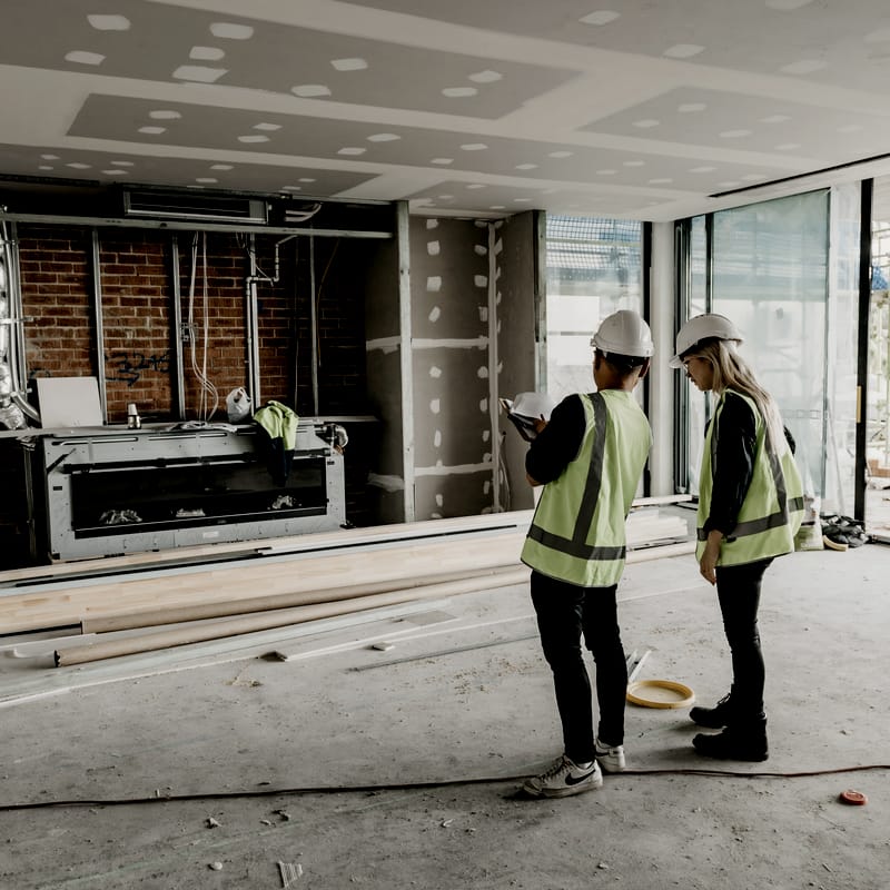Two people in hard hats and hi-vis vests reviewing plans inside a home under construction, with exposed brickwork and plasterboard walls