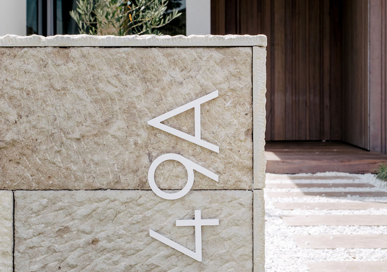 Close-up view of a stone entrance pillar with the number 49A displayed in white, modern font. In the background, there's a glimpse of a wooden door and a sleek, minimalist walkway with white pebble stones, reflecting one of the best duplex designs by a Sydney duplex architect.