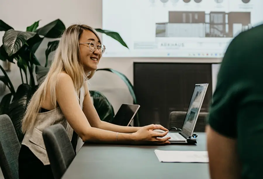 A person with long hair and glasses is sitting at a conference table with a laptop. They are smiling and facing another person who is partly visible. Large green plants adorn the background, while a projection screen displays a Sydney duplex architect's plans.